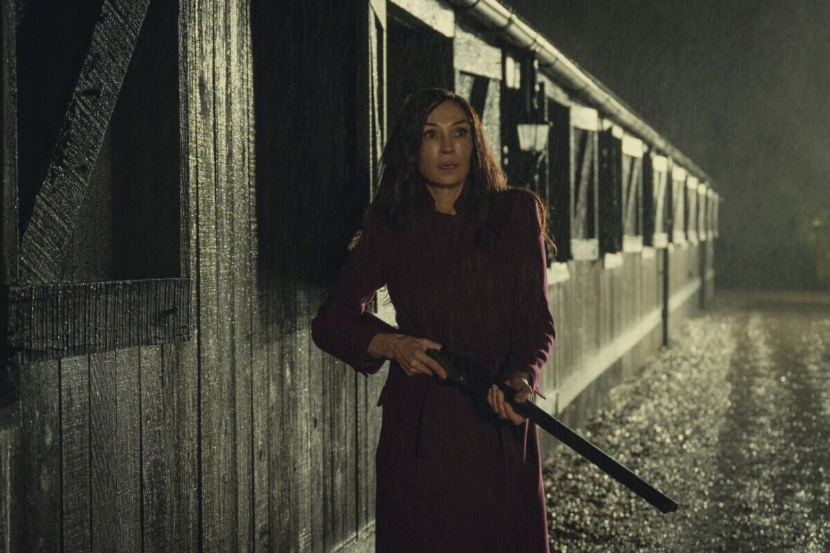 Za deštivého večera stojí žena u stájí s puškou v ruce.